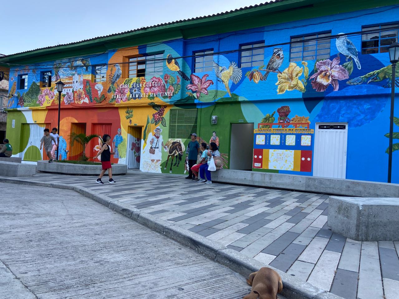 Fachada de la Casa de la Cultura Pablo A. Ramírez Martínez, en Nariño, con la intervención artística que la cubre de flores, aves y símbolos del municipio, y gente caminando por la plazoleta frente a ella.