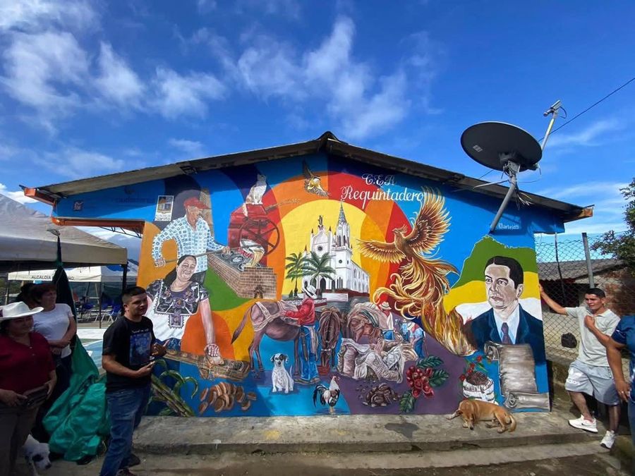 Mural del Centro Educativo Rural Requintadero, en la vereda El Roble, con escenas de la vida campesina, un ave fénix y vecinos junto a un perro frente a la fachada de la escuela.